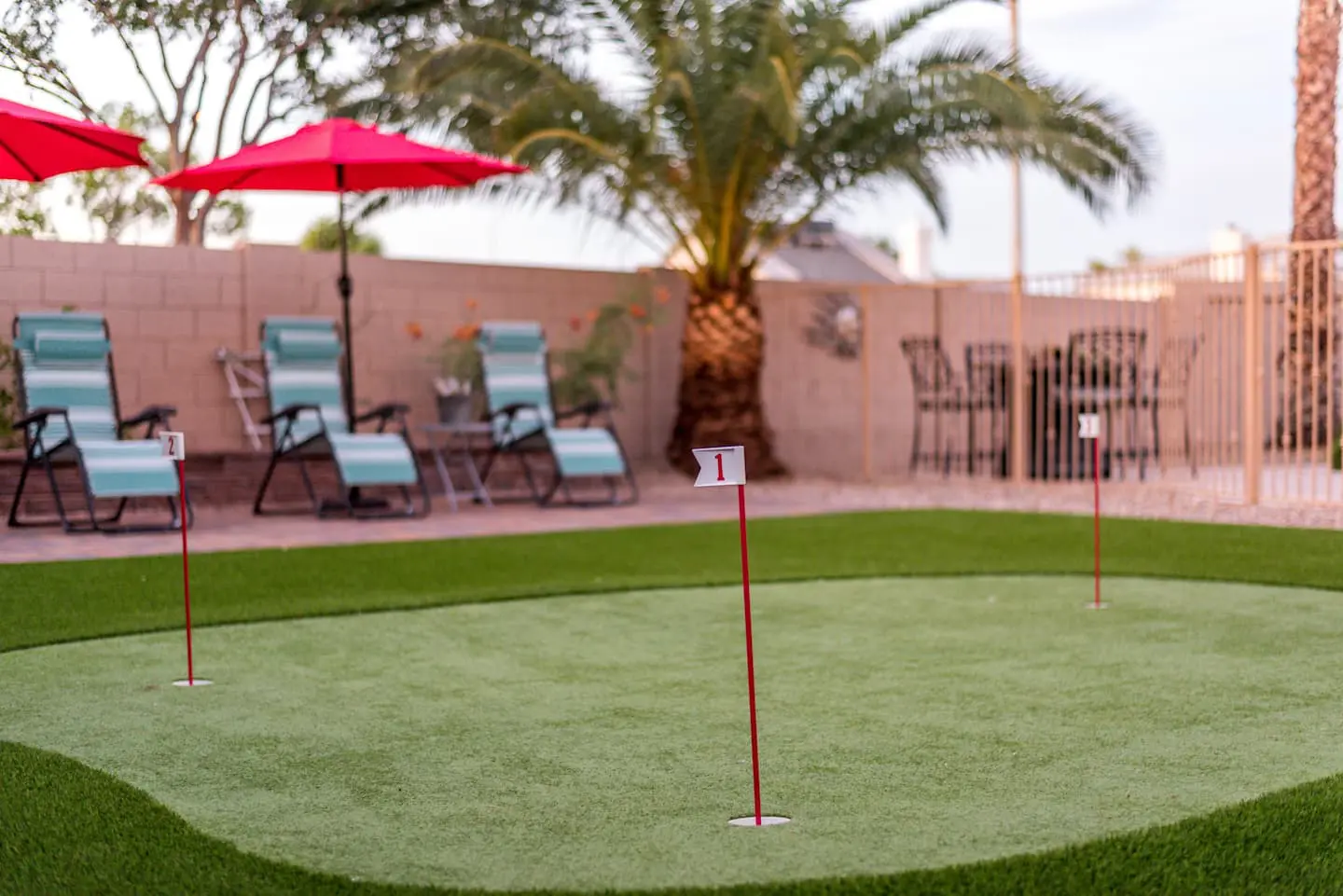 A small putting green with three red flag markers is in a backyard setting featuring pet-friendly turf. Lounge chairs sit under red umbrellas, and palm trees are visible in the background. A fence encloses the area, and a table with chairs is seen in the corner.