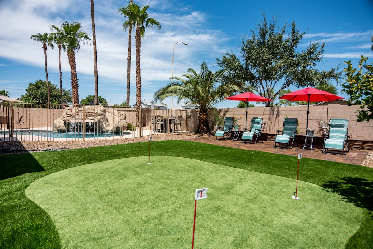 A backyard featuring a putting green with two holes, lush artificial grass, and a patio area with lounge chairs shaded by red umbrellas. There are tall palm trees and other greenery, and a waterfall feature near the back fence under a clear blue sky. This pet-friendly turf is perfect for family fun.