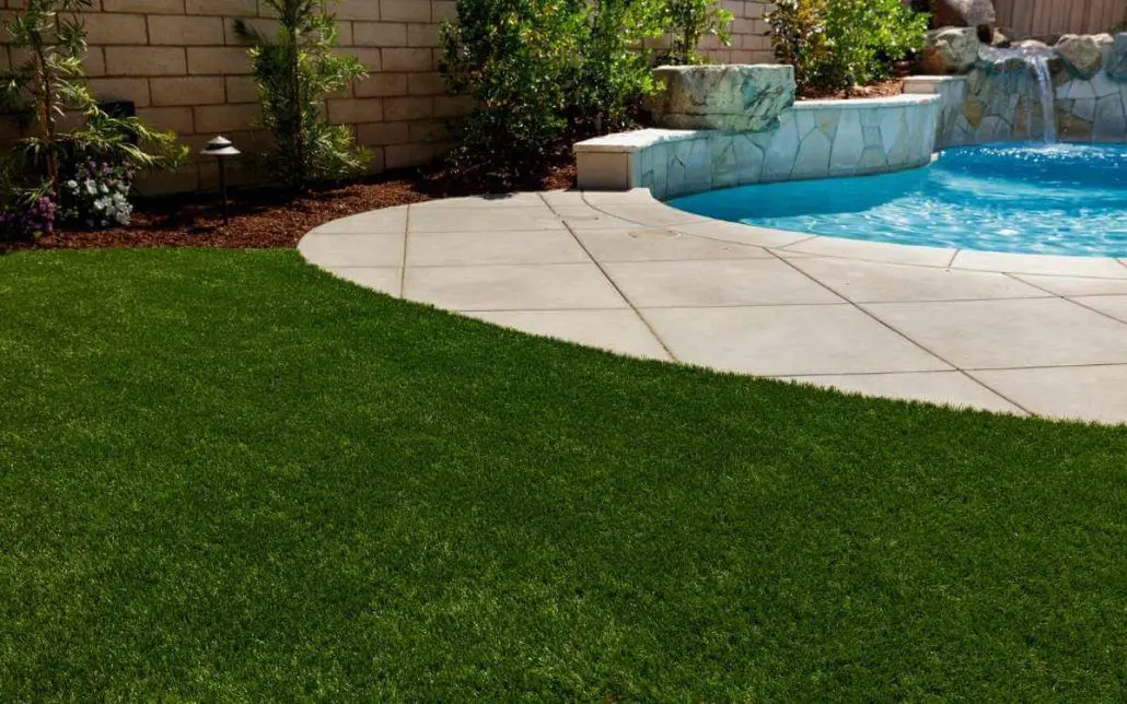 A backyard swimming pool with a stone waterfall feature, surrounded by a concrete pool deck that merges into well-manicured putting greens. The area is bordered by a stone wall with landscaping plants along its edge.