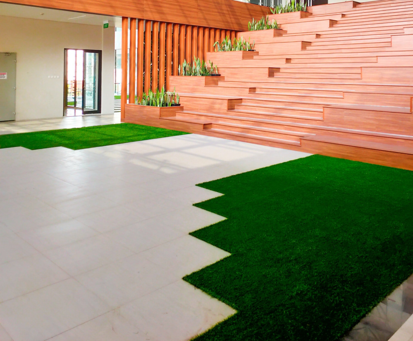A modern indoor space featuring a stepped wooden seating area adorned with potted plants on the steps. A mix of white tile flooring and pet-friendly turf fills the foreground. Sunlight streams through windows, creating a bright and inviting atmosphere.