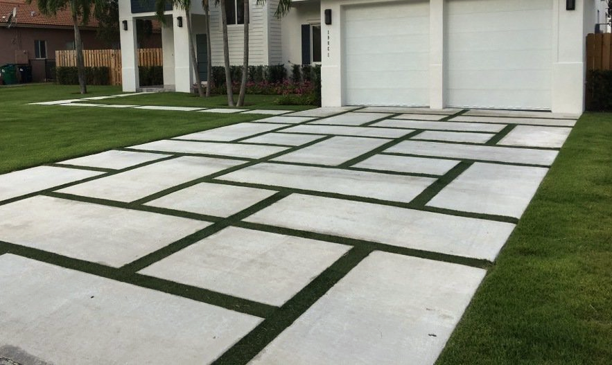 Modern driveway featuring large concrete slabs separated by narrow strips of artificial grass. The driveway leads up to a two-car garage attached to a contemporary white house. The surrounding lawn is well-maintained, and there are some trees and shrubs nearby.