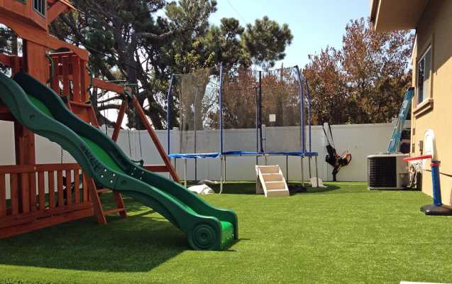 A backyard playground featuring a green slide, a wooden play structure, a large trampoline with a safety net, a swing, and a small rock climbing wall. The ground is covered with pet-friendly turf, and there is a beige house and white fence in the background.